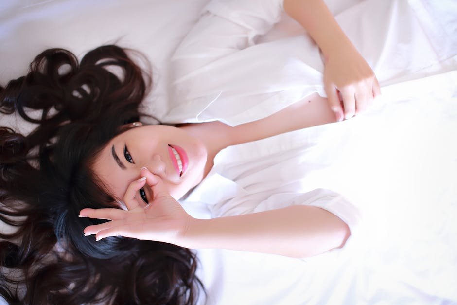 A cheerful woman lying on a bed, wearing a white shirt, smiling gently.