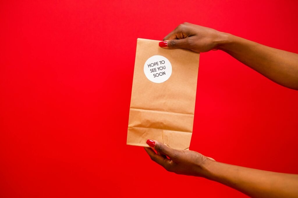 Two hands holding a paper bag with a 'Hope to see you soon' sticker, against a red background.